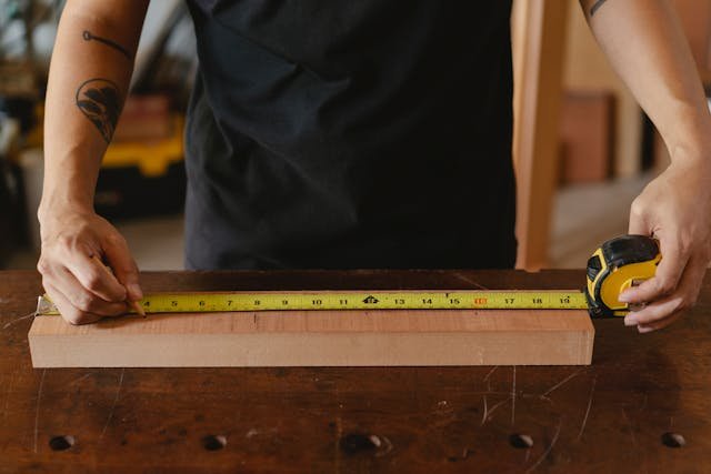 Person measuring a wooden plank with tape measure on workbench.