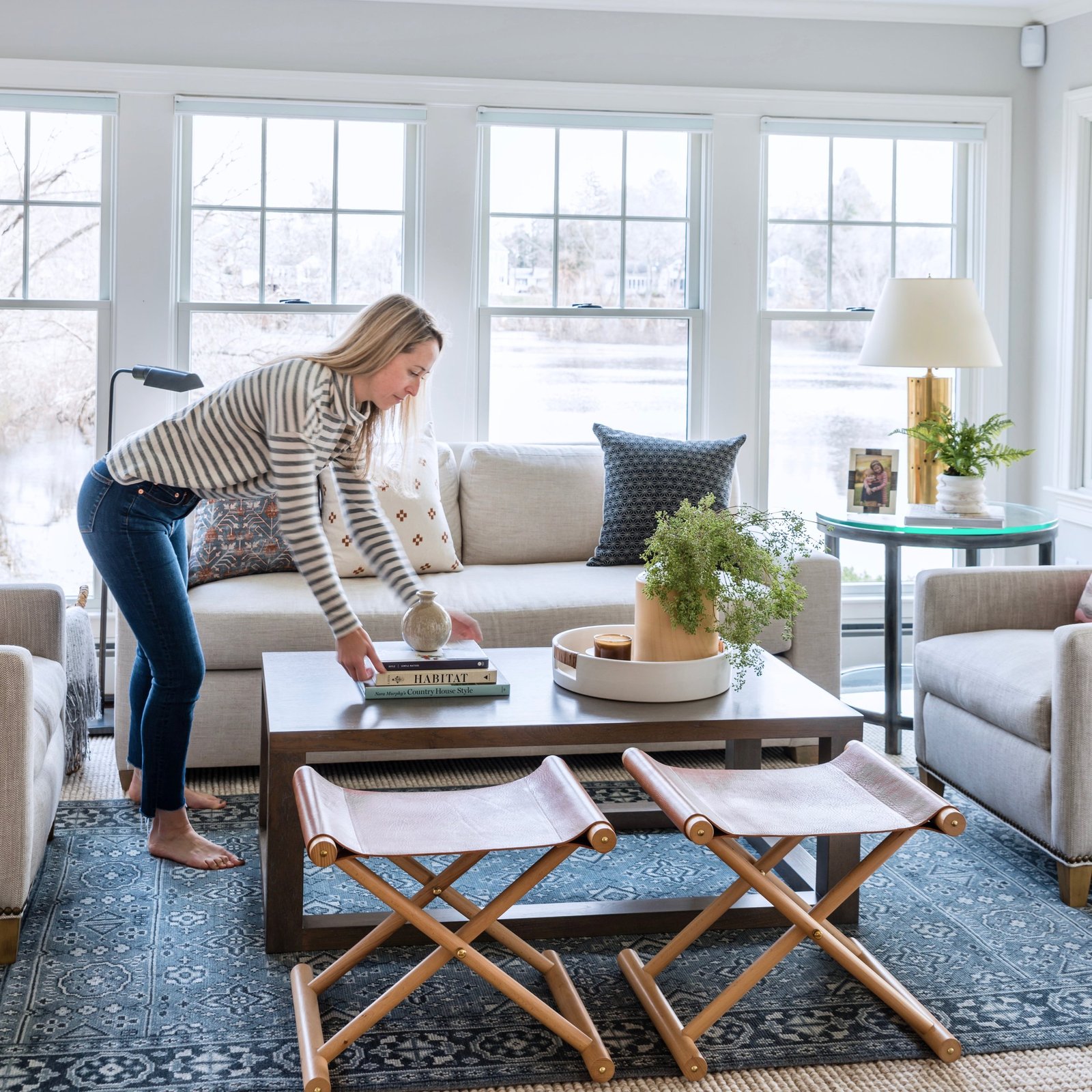 Woman arranging books on coffee table in a bright lakeside living room.