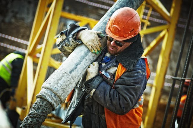 Construction worker in safety gear handling large concrete pouring hose.